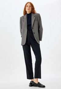 Grauer Hahnentrittmuster-Blazer mit strukturierter Schulterpartie, schwarzem Rollkragenpullover, taillierten schwarzen Hosen und schwarzen Loaferschuhen mit Fransenverzierung.