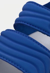 Μπλε σανδάλι με διαφάνειες που έχει ραβδωτή υφή, λείο υλικό και αντίθετες ραφές. Οι λουρίδες είναι φαρδιές και καμπυλωτές για άνεση.