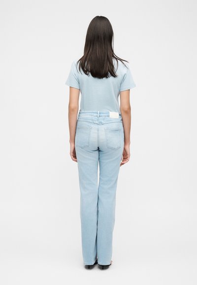 Γυναίκα με μακριά σκούρα μαλλιά, που φοράει ένα ανοιχτό μπλε μπλουζάκι και ανοιχτό μπλε τζιν, στέκεται με την πλάτη προς ένα λευκό φόντο.