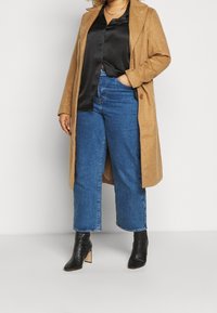 Abrigo de lana color canela, blusa de satén negra y jeans recortados de cintura alta azules, combinados con botas negras de tobillo con un tacón bajo.