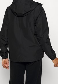 Chaqueta negra con capucha, con puños y dobladillo elásticos, fabricada en un tejido liso y waterproof, con un diseño simple y sin adornos.