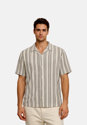 Jonge man die een kort mouw overhemd met verticale zwart-witte strepen draagt, staand tegen een effen achtergrond.