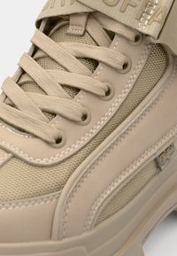 Beige sneaker met gestructureerde mesh panels, platte veters en contrasterende stiksels; heeft een opvallend logo op de band en een rubberen zool.