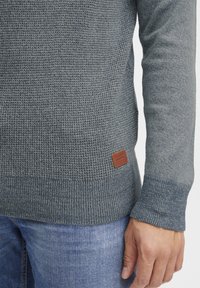Knitted sweater in dark blue-gray, featuring a textured pattern. Includes a brown leather tag. Cuffs and hem are ribbed for detail.