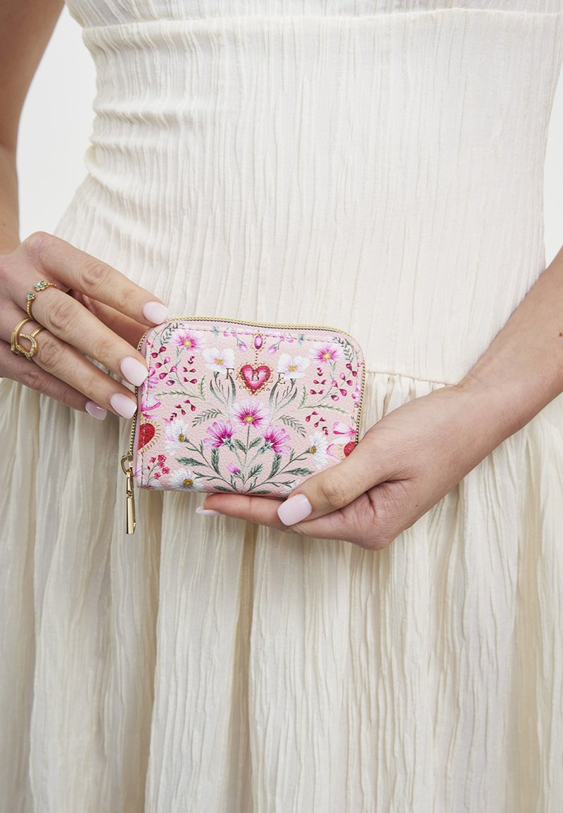 Mains tenant un petit portefeuille rose avec broderie florale et fermeture éclair dorée, porté sur une robe blanche texturée.