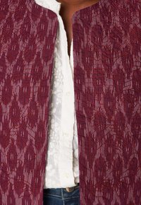 Primo piano di una persona che indossa una camicia bianca ricamata con bottoni sotto una giacca bordeaux a pattern con strisce verticali e fantasie floreali.