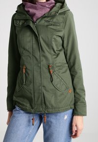 Veste à capuche vert olive avec cordons, deux poches avant et une fermeture éclair, associée à une écharpe violette et un jean en denim.