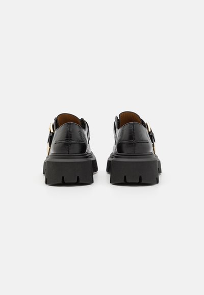 Mocassini in pelle nera con texture in coccodrillo, suole chunky lug e dettagli in oro, mostrati da dietro su uno sfondo bianco.