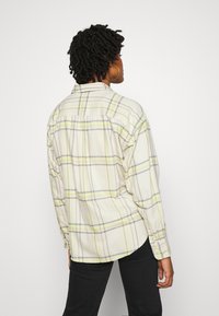 Person with short curly hair wearing hoop earrings and a loose cream plaid shirt, facing away, paired with black pants.