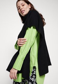 Svart strikket poncho med høy hals, lagdelt over en levende grønn skjorte med knappest closure. Merkbare sidesplitter og kontrasterende teksturer.
