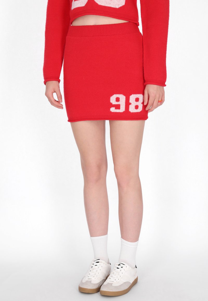 Röd stickad kjol med en åtsittande midja, som visar en vit "98" på vänster sida. Kombinerad med vita strumpor och beige sneakers.