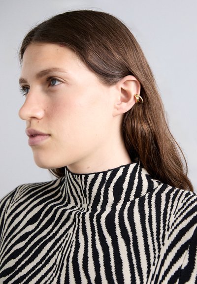 Guldøre cuff båret på den øverste del af øret med en sort- og cremefarvet stribet højhalset sweater. Glat tekstur og dristige kontrasterende mønstre.