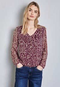 Doorzichtige bordeauxrode blouse met een v-hals en lange, opgezette mouwen. Kenmerkt zich door een abstract patroon en geplooide details bij de manchetten. Gecombineerd met denim.