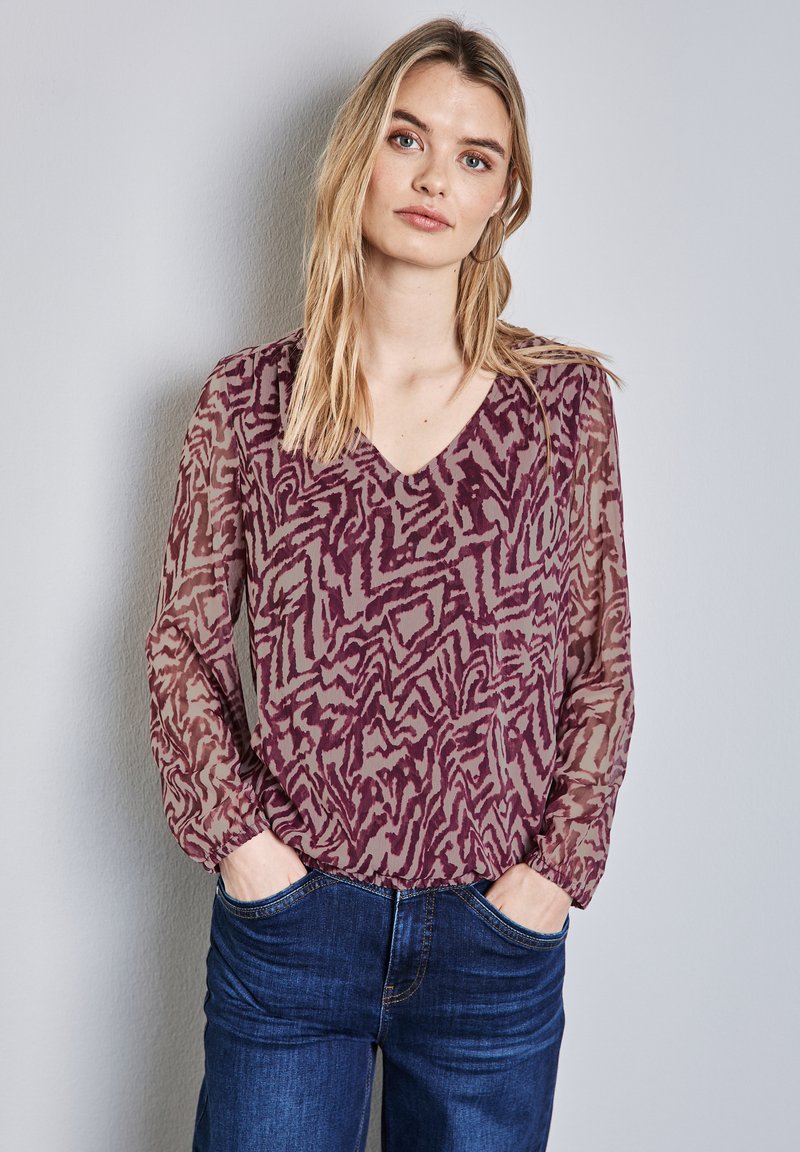 Doorzichtige bordeauxrode blouse met een v-hals en lange, opgezette mouwen. Kenmerkt zich door een abstract patroon en geplooide details bij de manchetten. Gecombineerd met denim.
