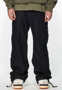 Black cargo pants with multiple side pockets, made from durable fabric. The cuffs are adjustable. Worn with light-colored sneakers.
