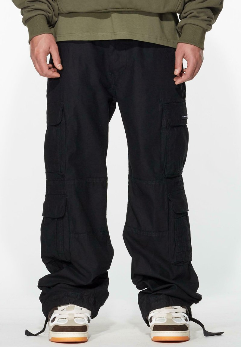 Black cargo pants with multiple side pockets, made from durable fabric. The cuffs are adjustable. Worn with light-colored sneakers.