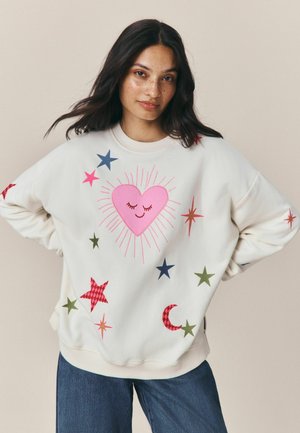 Junge Frau mit langen dunklen Haaren, die einen weißen Pullover trägt, der mit bunten Sternen, einem rosa lächelnden Herz und einem Halbmond verziert ist.