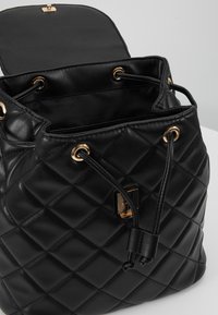 Mustakuvioinen nahkainen naruhandbag, jossa on kultasävyiset metalliset rei'itykset ja salpa, avoin läppä, joka näyttää sisävuoren.