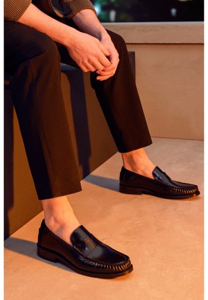 Personne assise les mains jointes, portant un pantalon noir et des mocassins en cuir noir brillant sans chaussettes sur un sol beige.