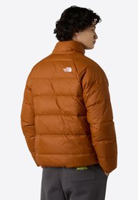 Orange pufferjakke med horisontale syninger, høj krave og hvidt logo på ryggen. Glat, skinnende overflade; tætsiddende pasform.