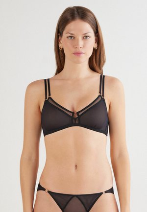 Woman with long brown hair wearing black sheer bra and matching underwear, standing against plain light background.