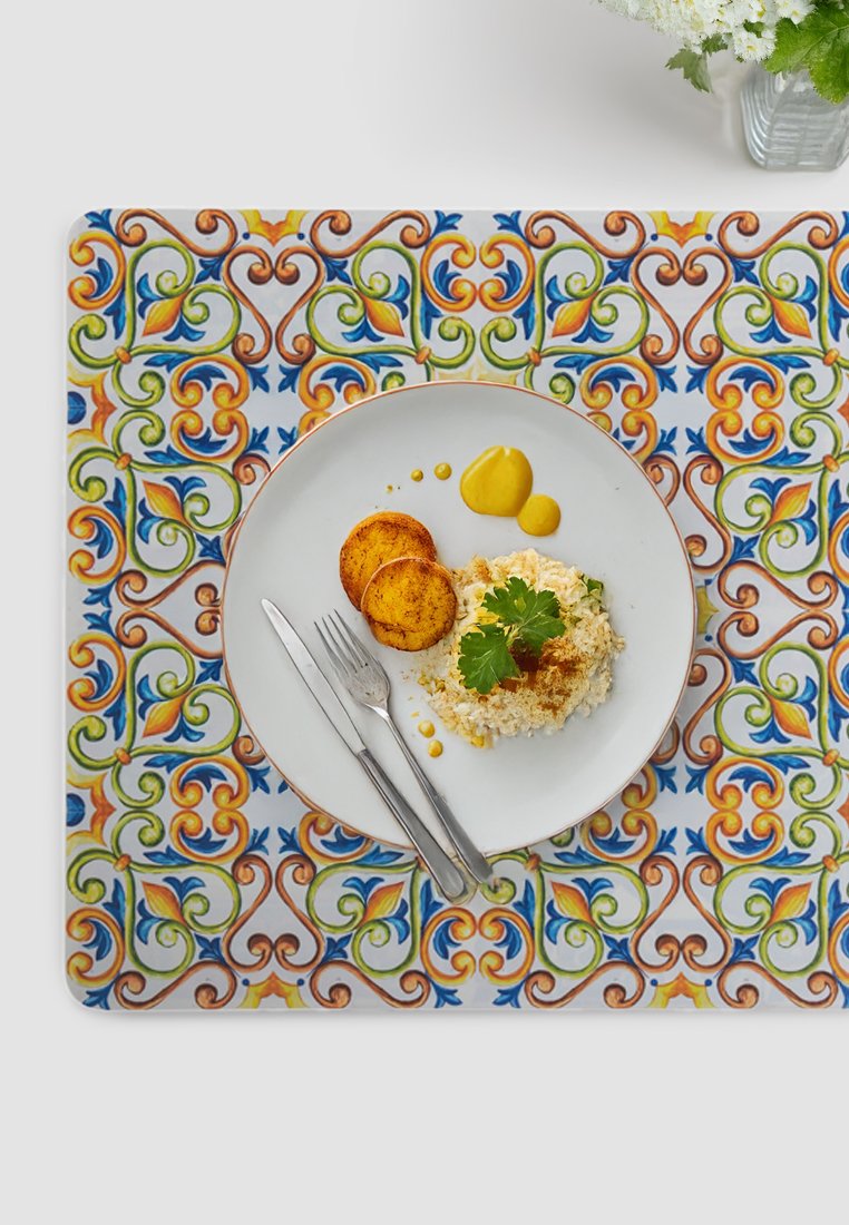 Kolorowy dekoracyjny podkładka na stół z misternymi wzorami w odcieniach pomarańczowym, niebieskim i zielonym. Na białym talerzu znajduje się ryż, dwa złote ziemniaki oraz sosy.