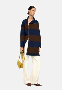 Paski, zapinany na zamek sweter w ciemnoniebieskim i brązowym kolorze, z wysokim kołnierzem i bocznymi rozporkami, zestawiony z kremowymi spodniami o szerokich nogawkach i żółtą torebką.
