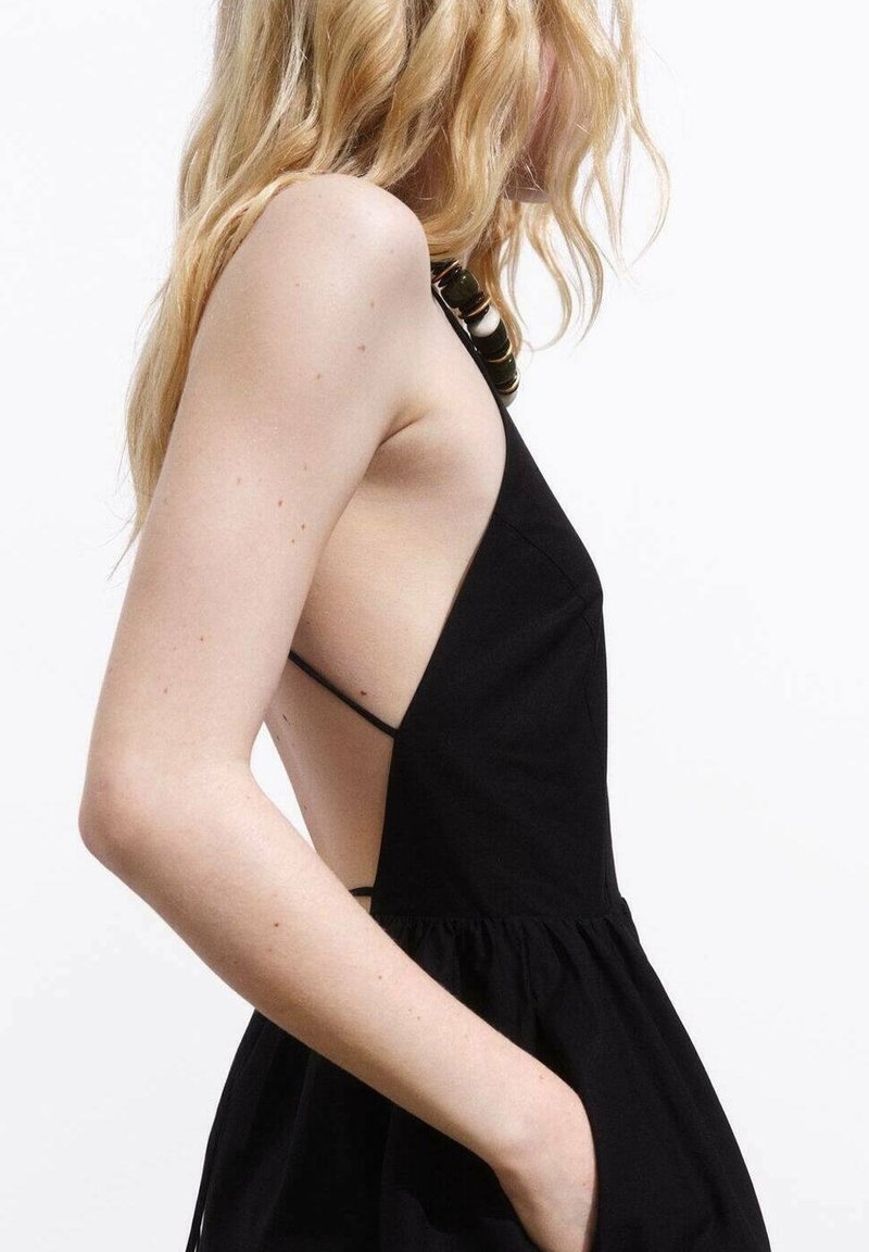 Femme blonde en robe noire sans manches avec côtés ouverts, posant avec une main dans la poche sur un fond blanc uni.