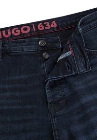 Jeans de mezclilla de lavado oscuro con una cinturilla de logo rojo, que presenta dos botones plateados y un acabado limpio en los bolsillos y costuras.