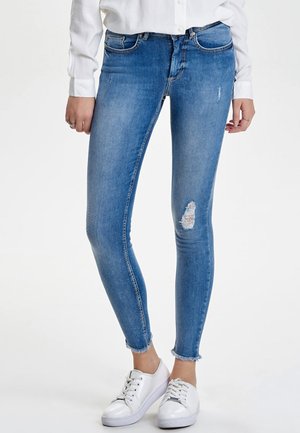 Jean skinny en denim bleu clair avec un genou déchiré, des ourlets effilochés et un design à cinq poches, associés à des baskets blanches.
