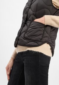 Gestepptes schwarzes Weste mit einer Reißverschlusstasche, runder Ausschnitt und abgerundetem Saum. Getragen über einem beigen Strickpullover und dunklen Jeans.