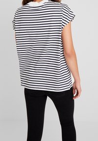 T-shirt a righe bianco e navy, maniche corte, vestibilità ampia, scollatura rotonda, con orlo leggermente ribassato. Indossato con jeans neri.