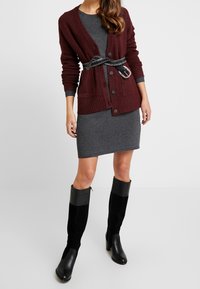 Burgundowa dziana kardigan na szarej sukience, zestawiona z czarno-szarymi kozaczkami na obcasie do kolan. Posiada kieszenie oraz pasek z srebrną klamrą.
