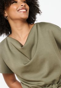 Olivno zelena draped majica s kratkimi rokavi, gladka tekstura in izrez s coto. Vključuje detajl z vezavo v pasu.