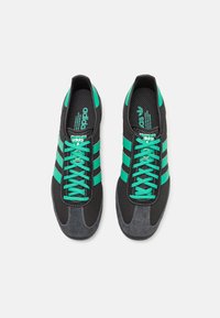 Sapatos desportivos pretos com detalhes verdes, apresentando um cabedal em malha, design com três riscas, atacadores e uma biqueira em camurça cinza.