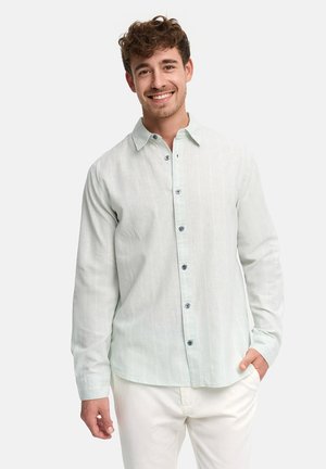 Homme souriant aux cheveux bouclés portant une chemise boutonnée vert clair et un pantalon blanc, debout avec une main dans la poche sur fond blanc.