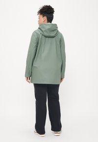 Πράσινο αδιάβροχο μπουφάν με κουκούλα, μακριά μανίκια και χαλαρή εφαρμογή, συνδυασμένο με μαύρο φαρδύ παντελόνι και αθλητικά παπούτσια ανοιχτού χρώματος.