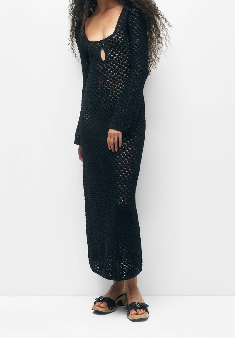 Femme portant une longue robe noire en maille ajourée avec manches longues et décolleté plongeant, associée à des sandales noires à talons avec nœuds.