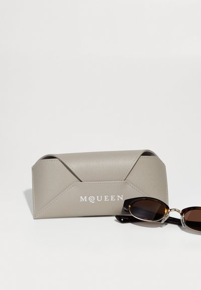 Husa de ochelari McQueen din piele gri, cu ochelari ovali tortoisefoldați lângă ea, pe un fundal alb.