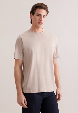 Jonge man die een beige T-shirt met korte mouwen en donkere broek draagt, staand tegen een effen lichte achtergrond, recht vooruit kijkend.