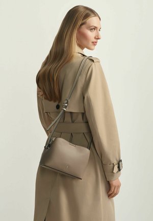 Kobieta o długich blond włosach, ubrana w beżowy płaszcz przeciwdeszczowy, nosząca taupe skórzaną torbę na ramię, na jednolitym jasnym tle.