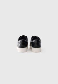 Et par svarte lær sneakers med hvite såler, sett fra baksiden, på en ensfarget hvit bakgrunn.