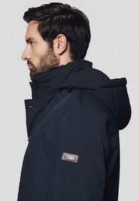 Granatowa kurtka ocieplana z wysokim kołnierzem i regulowanym kapturem. Posiada pikowaną strukturę oraz naszywkę z logo na rękawie.