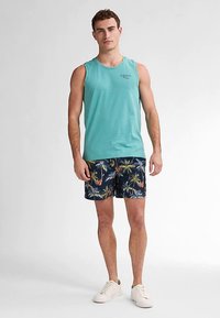 Vaaleanpetrolinvihreä hihaton paita, jossa on logo, yhdistetty tummansinisiin kukkakuviollisiin uimashortseihin. Malli käyttää valkoisia lenkkitossuja. Neutraali tausta.