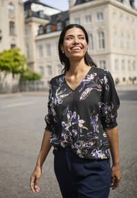 Donna sorridente che cammina all'aperto indossando una blusa floreale nera e pantaloni blu scuri, con edifici storici sullo sfondo.