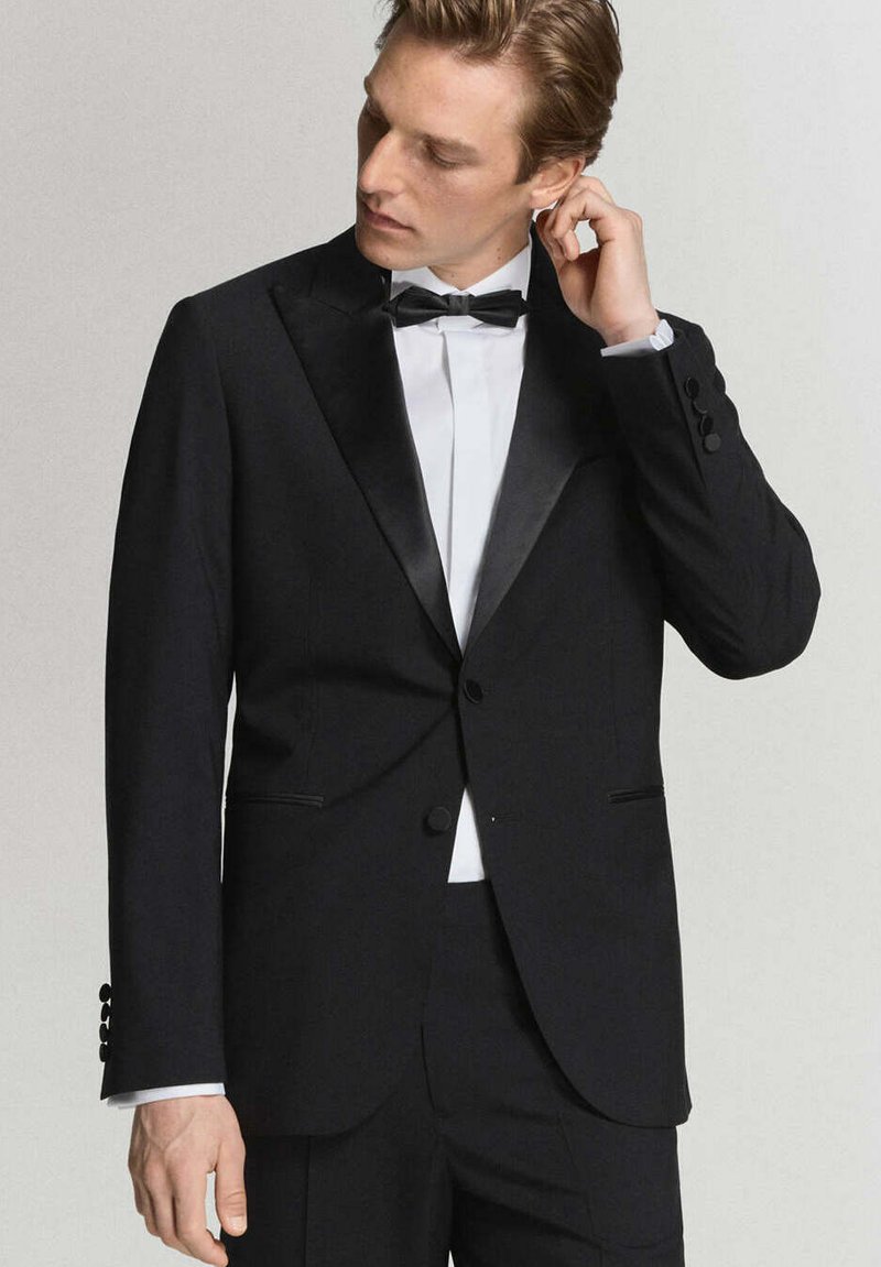 Hombre ajustándose la oreja, vistiendo chaqueta de esmoquin negra con solapas de satén, camisa blanca y pajarita negra sobre fondo liso.