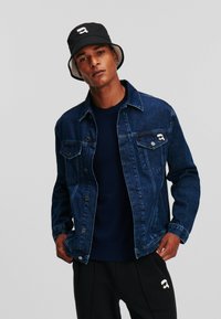 Denim jack in donkerblauw met voorzakken en zilverkleurige hardware, gelaagd over een marineblauwe trui. Zwarte bucket hat met een logo.