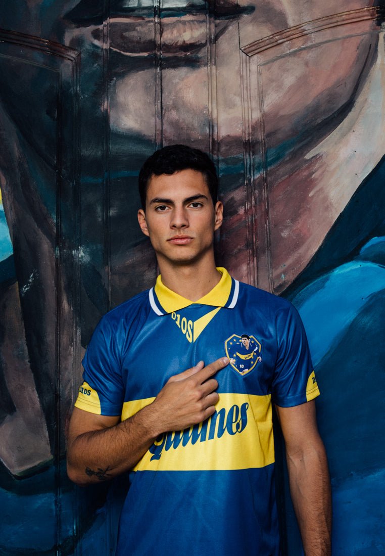 Blauwe voetbalshirt met gele accenten, heeft een embleem op de borst, korte mouwen en een kraag met gestreepte details.