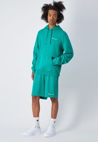 Türkiser Sweatshirt mit Kapuze und Kängurutasche, kombiniert mit passenden Shorts. Beide zeigen das Champion-Logo und werden mit weißen Sneakers und Socken getragen.