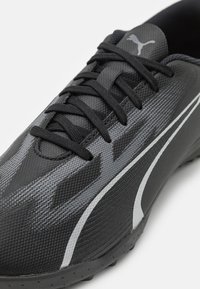 Czarne buty sportowe o teksturowanym materiale, z szarymi akcentami i kontrastowym białym logo. Widać sznurówki i gumową podeszwę.